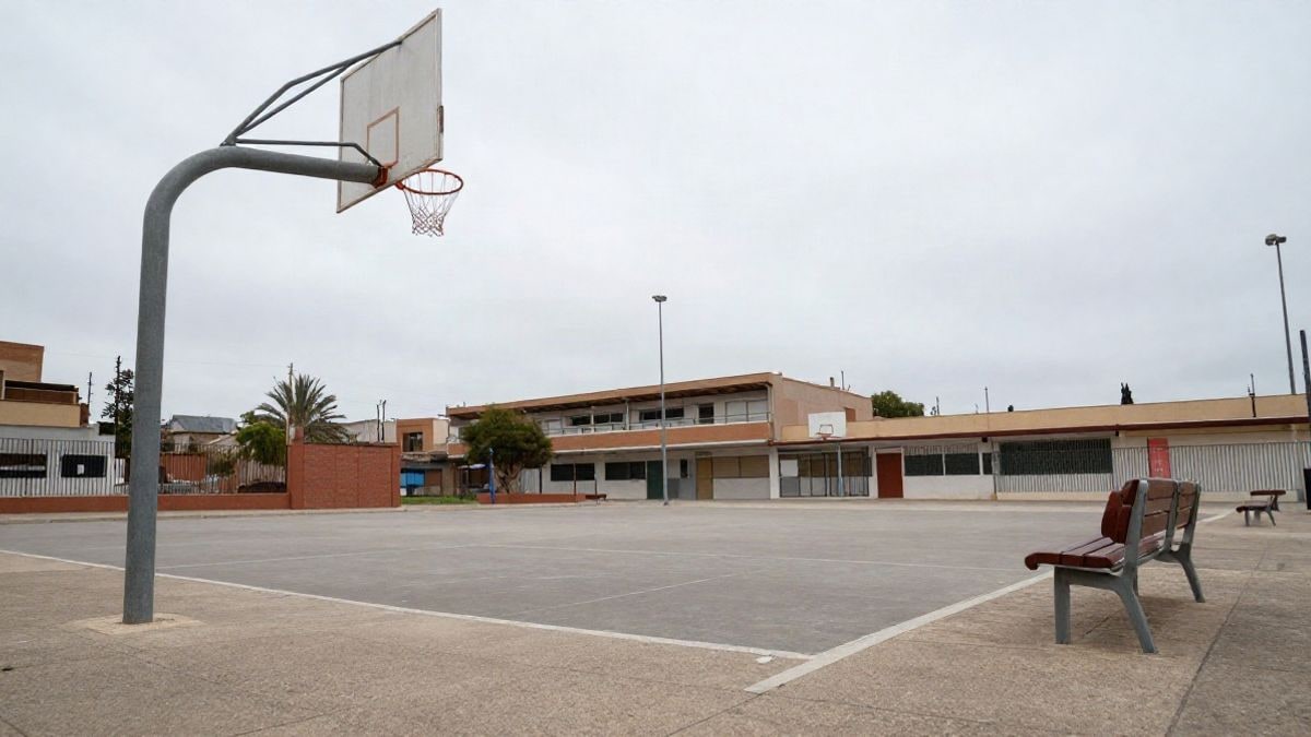 Imagen ilustrativa: ángulo bajo de un patio escolar vacío con canasta y banco bajo un cielo nublado