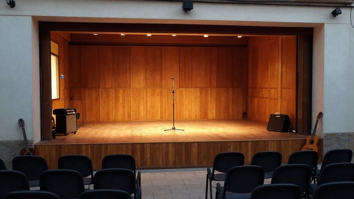 Imagen ilustrativa: interior de una sala de conciertos con escenario de madera y un micrófono, con luz lateral de ventana