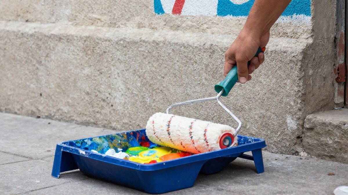 Imagen ilustrativa: primer plano de un rodillo de pintura y una bandeja con pintura de colores junto a un muro de hormigón