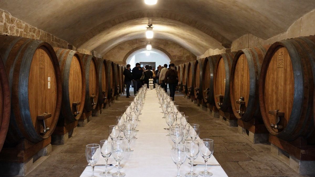 Imagen ilustrativa: interior de una bodega con barricas de madera y una mesa larga con copas de vino vacías.