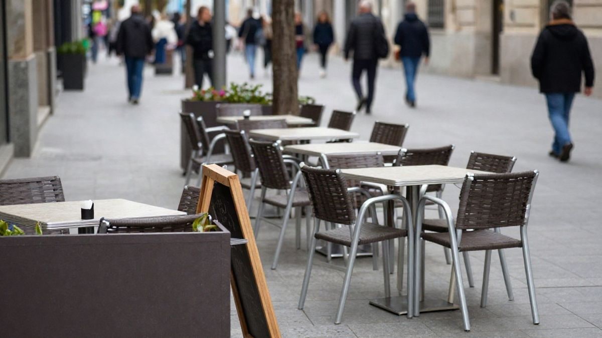 Imagen ilustrativa: sillas y mesas vacías de una terraza en un paseo urbano, con una pizarra y un separador de plantas