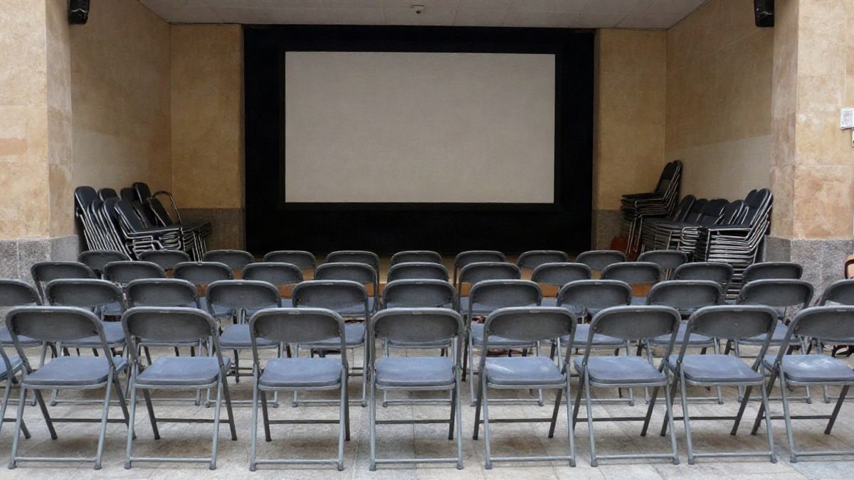 Imagen ilustrativa: filas de sillas plegables vacías ante una pantalla de proyección en una sala cultural municipal