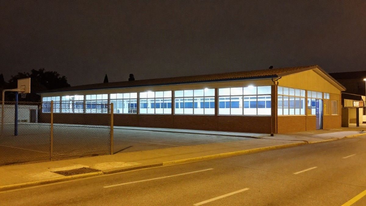 Imagen ilustrativa: edificio anexo de un gimnasio escolar iluminado de noche junto a un patio con canasta