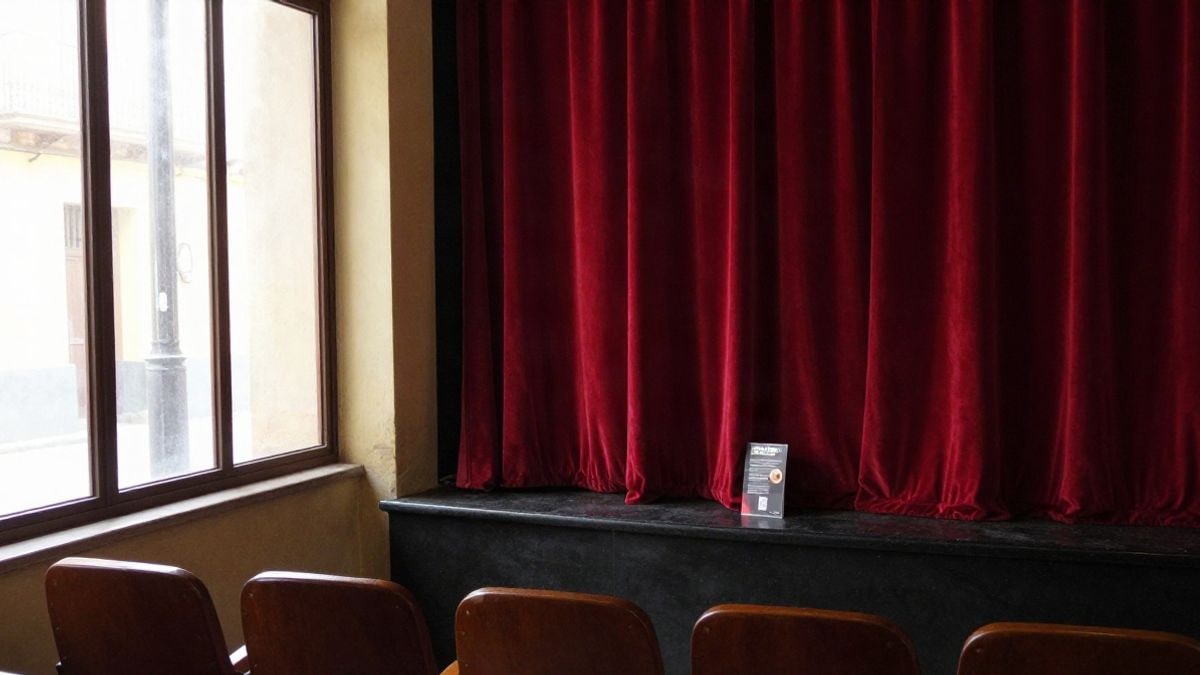 Imagen ilustrativa: escena de interior de un teatro con cortinas rojas y butacas vacías junto a una ventana lateral