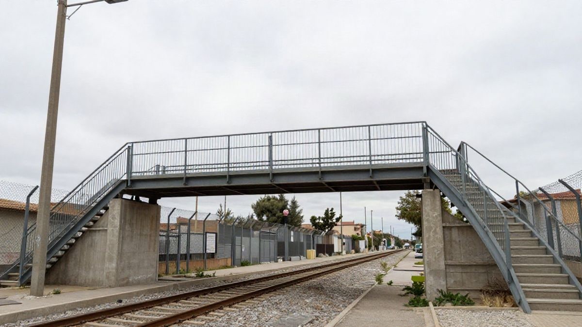 Imagen ilustrativa: pasarela metálica con escaleras y rampa sobre vías de tren en un entorno urbano