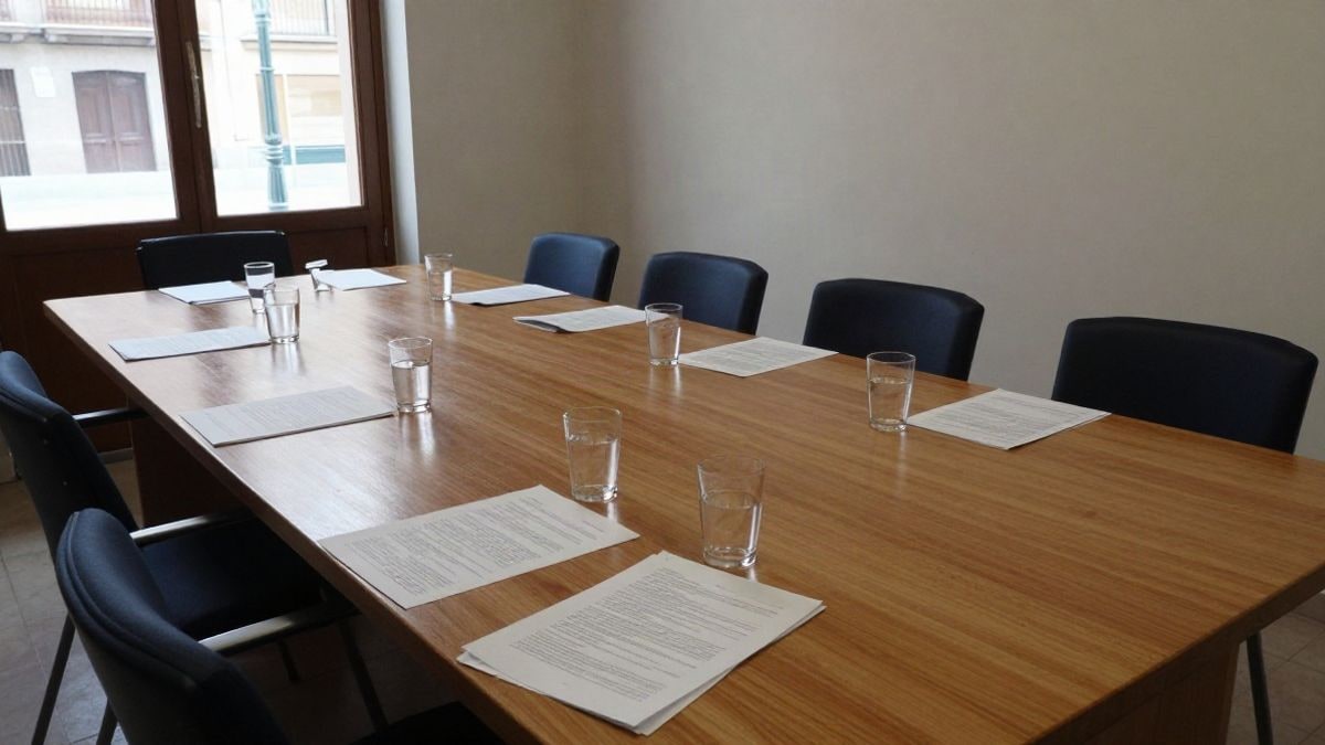 Imagen ilustrativa: sala de reuniones municipal con mesa larga y sillas vacías junto a una ventana