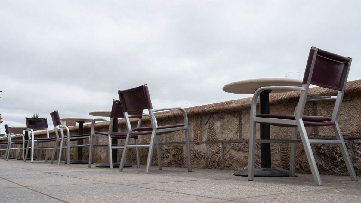 Imagen ilustrativa: sillas y mesas vacías de una terraza en una calle corriente, con cielo nublado de fondo
