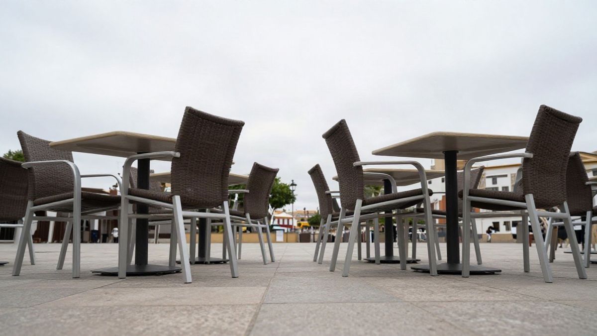 Imagen ilustrativa: mesas y sillas vacías en una plaza pavimentada bajo un cielo nublado