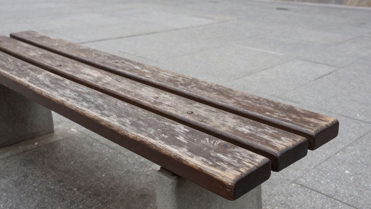 Imagen ilustrativa: superficie de madera envejecida de un banco en una plaza pública con baldosas grises