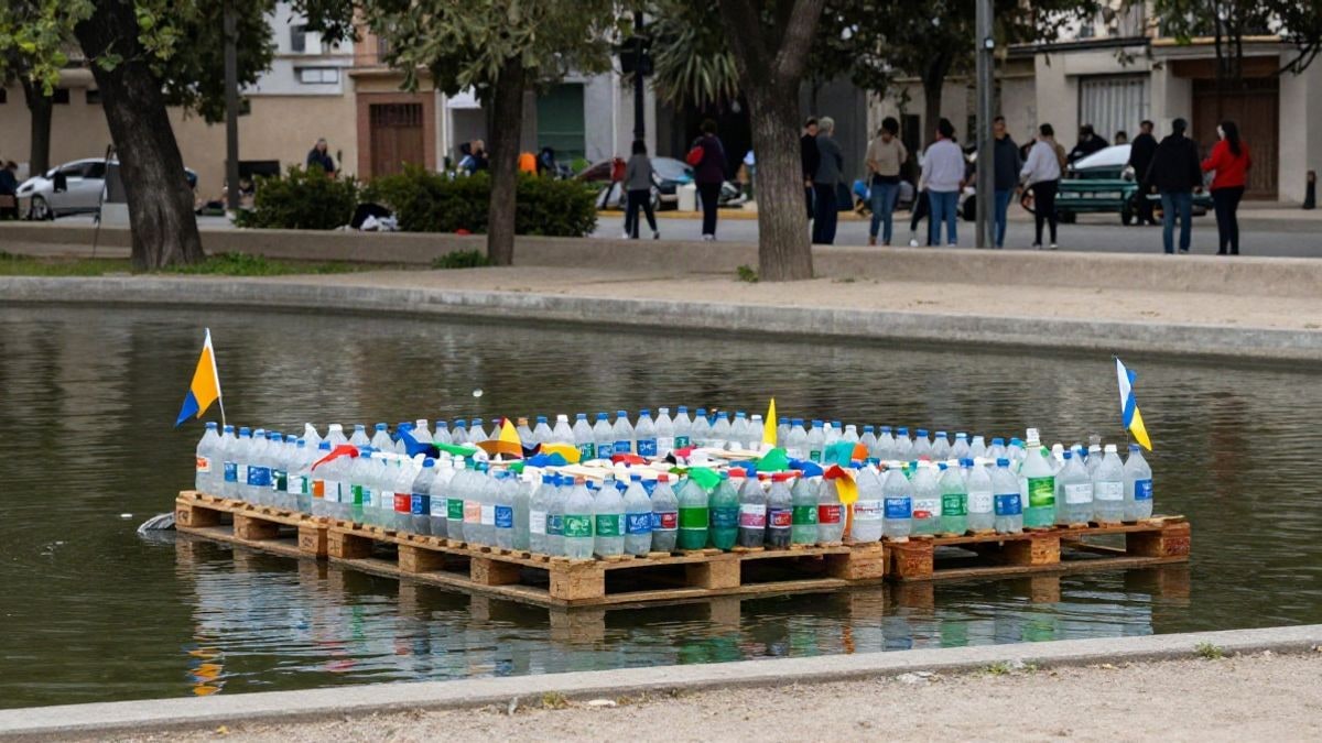 Imagen ilustrativa: una balsa casera hecha con botellas de plástico y palés flotando en un estanque de un parque urbano, con banderines de colores.