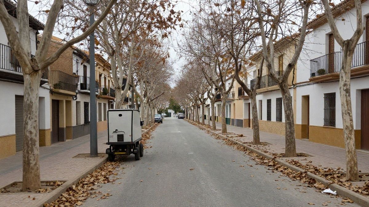 Imagen ilustrativa: calle arbolada de un pueblo mediterráneo con hojas caídas en la acera y un carro de limpieza aparcado en un día nublado